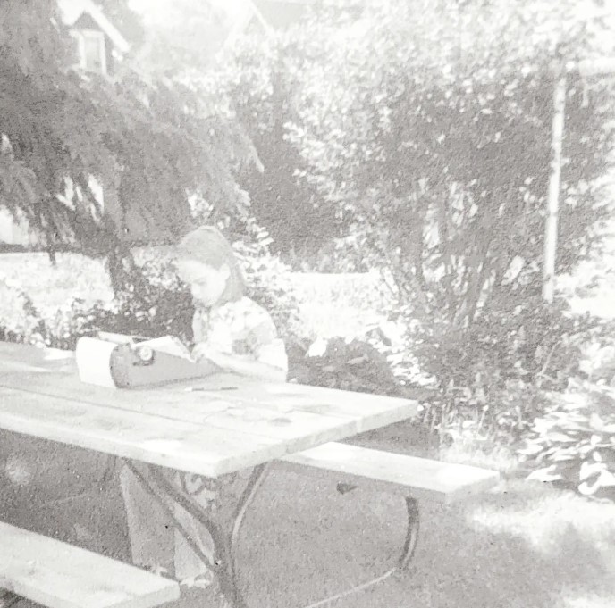 black and white photograph of straight blond haired young child sitting at a back yard picnic table using a manual typewriter with leafy trees in the background