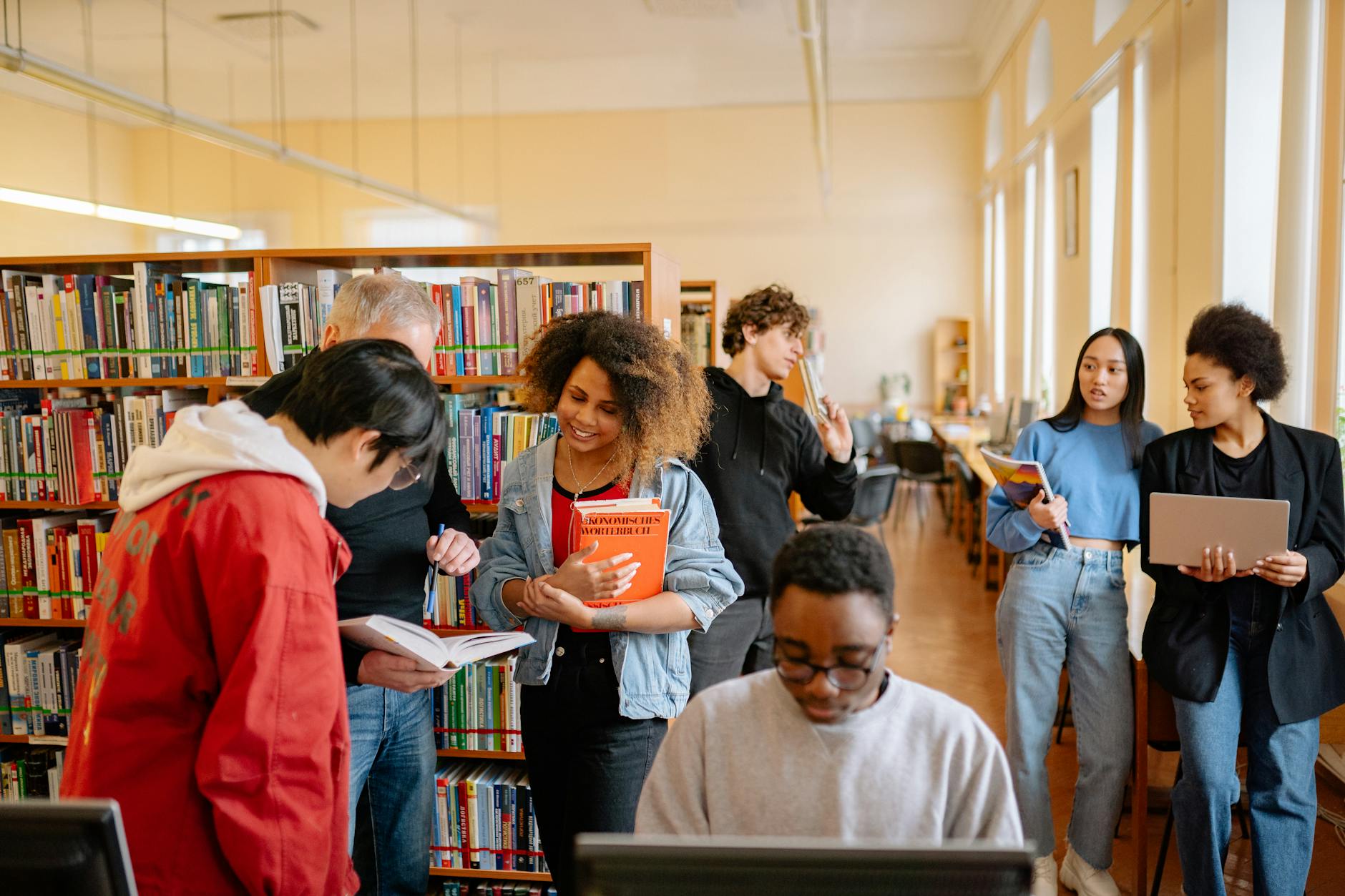 7 different young college age folks of different backgrounds talking looking at books or laptops inside a library 